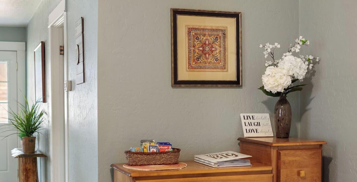 Un couloir avec un banc en bois, une œuvre d’art encadrée, un panier rempli d’objets, un vase avec des fleurs et un panneau d’affichage avec un message motivant.