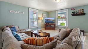 Ein Wohnzimmer mit einem großen, beigefarbenen Ecksofa, einem Holztisch und einem Fernseher auf einem Schrank. Es gibt zwei Fenster mit Jalousien, eine Lampe und ein Schild an der Wand.