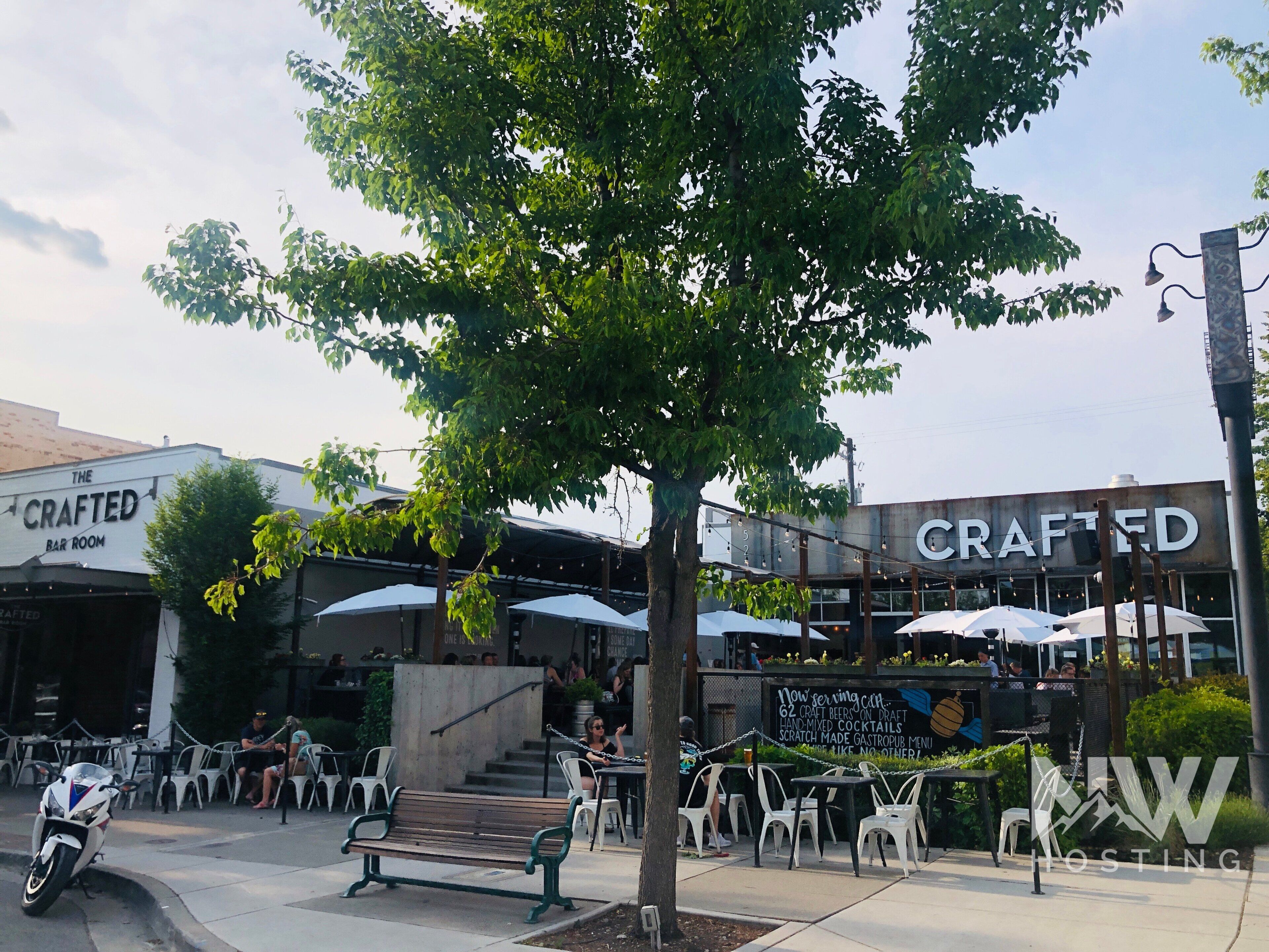 An outdoor seating area with tables and chairs, a tree, and a building with 'CRAFTED' signage.