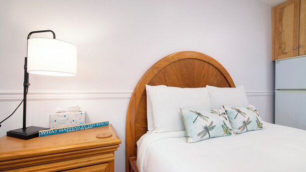 A neatly made bed with a wooden headboard, a bedside lamp, a book titled "What Happens at the Beach," and a tissue box on a wooden nightstand. - Walkout Beachfront Condo: Pointes North Resort 106 (Traverse City)