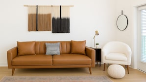 Una sala de estar moderna con un sofá de cuero marrón, un sillón blanco, un tapete estampado y una obra de arte abstracta colgada en la pared.