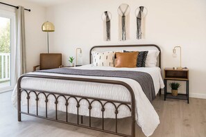 Un dormitorio con estructura de cama metálica, una mesita de noche con lámpara, una decoración de macramé en la pared y vistas al exterior.
