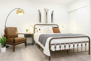 Un dormitorio con estructura de cama metálica, una butaca de cuero marrón, una mesita de noche con lámpara y decoración en la pared.