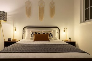 Una cama bien tendida con una colcha estampada, dos lámparas de noche y adornos de macramé colgados en la pared.