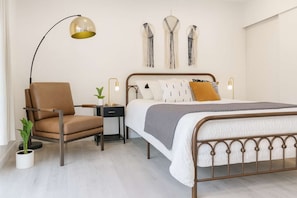 Un dormitorio con cama, una silla, mesita de noche, lámpara, decoración en la pared y una planta en maceta.