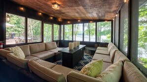 A spacious room with a large beige sectional sofa, a black coffee table, and a view of greenery through large windows. - Lakefront Hot Tub Canoes Game Room Wood FP (Gouldsboro)