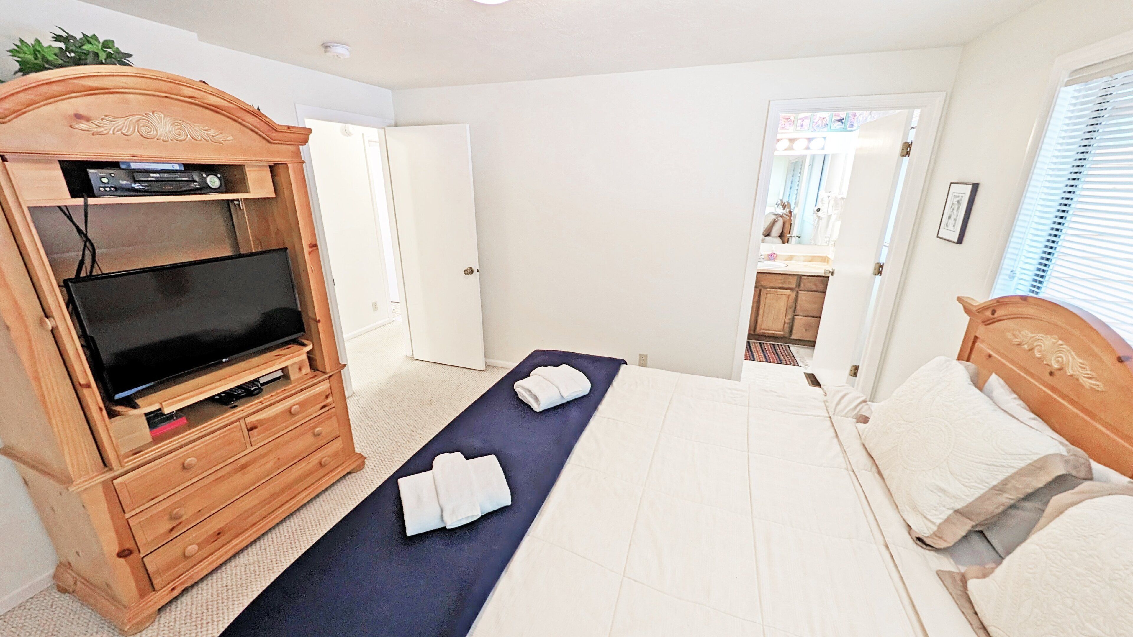 A bedroom with a TV stand, a bed, a blue mat, and a bathroom visible through an open door.
