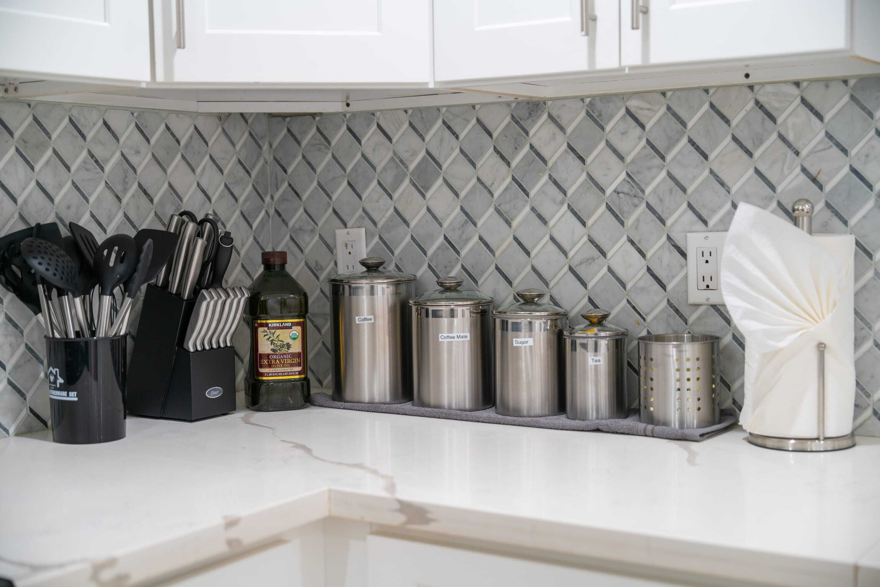 A kitchen with white cabinets, a diamond-patterned backsplash, and stainless steel containers labeled for coffee, coffee mate, and sugar.