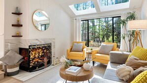 A modern living room with a fireplace, yellow armchairs, a round coffee table, and large windows with a view of greenery.