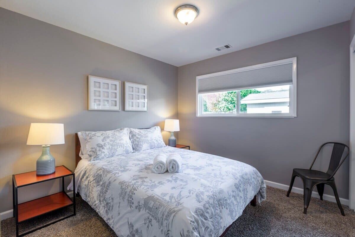 A bedroom with a bed, two bedside tables with lamps, a chair, a window, and a wall-mounted light fixture.