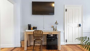 A small, neatly organized room with a wooden desk, a chair, a television mounted on the wall, a small refrigerator, and a white door. - The Royce on Spring - High-end Family Getaway in Newport (Newport)