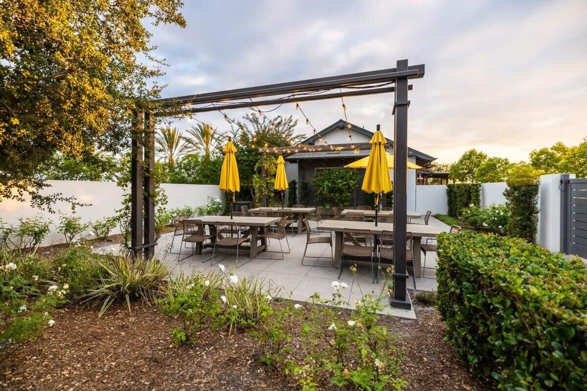 An outdoor patio with tables and chairs, a pergola with yellow umbrellas, and a building in the background.