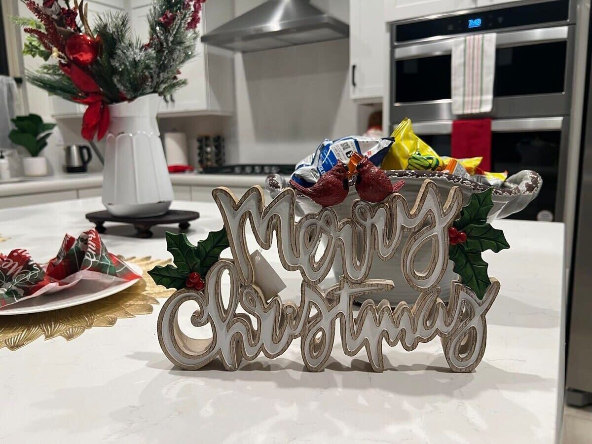 Una encimera de cocina con un cartel decorativo que dice 'Merry Christmas', un jarrón blanco con follaje y alimentos con motivos navideños.