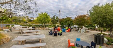 An outdoor seating area with picnic tables and chairs, surrounded by greenery and trees.