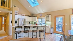 A kitchen with a central island, bar stools, a clock, and a view through the window. - Mcnary 20 (Bend)