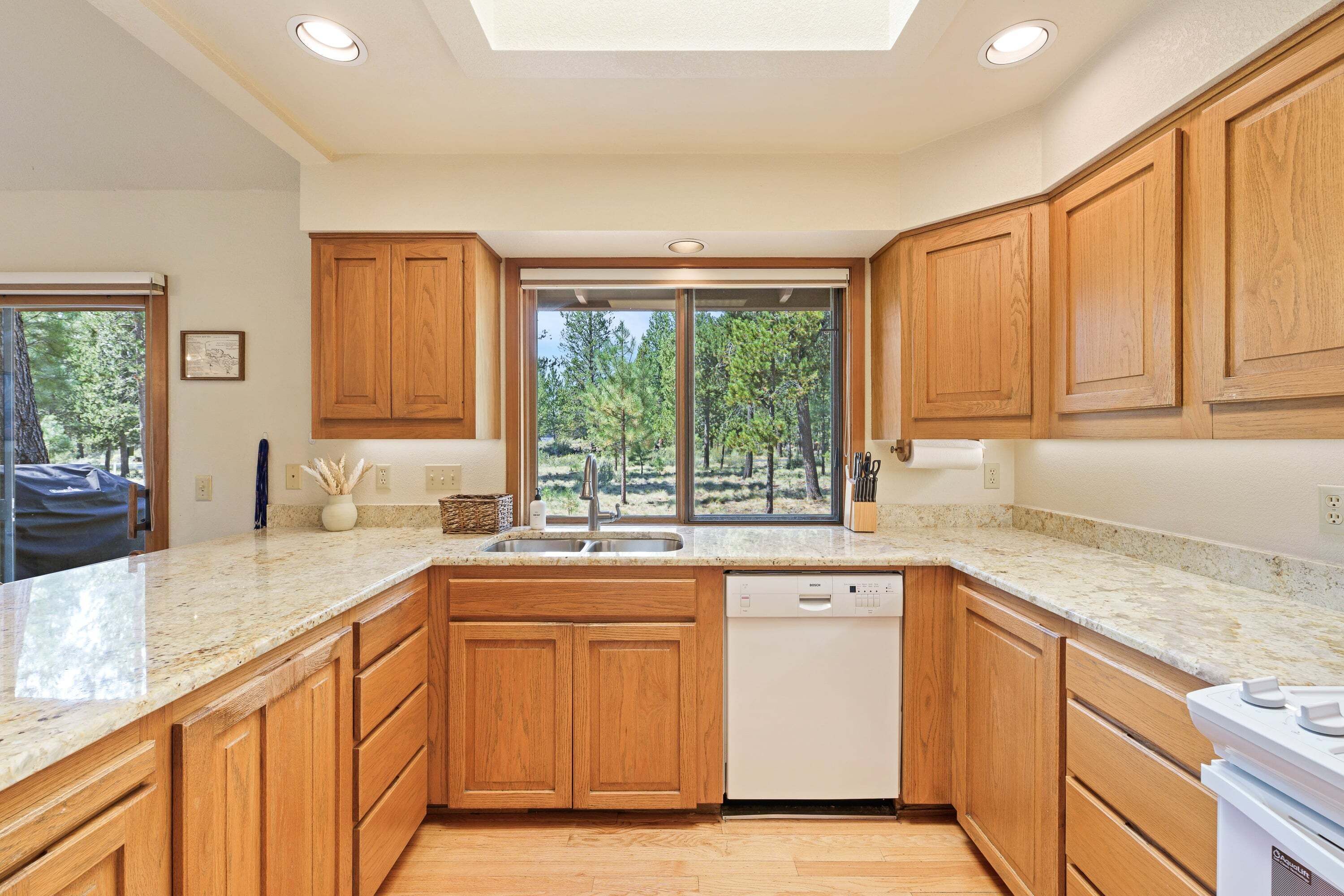 A modern kitchen with wooden cabinets, a dishwasher, and a large window with a view of trees.