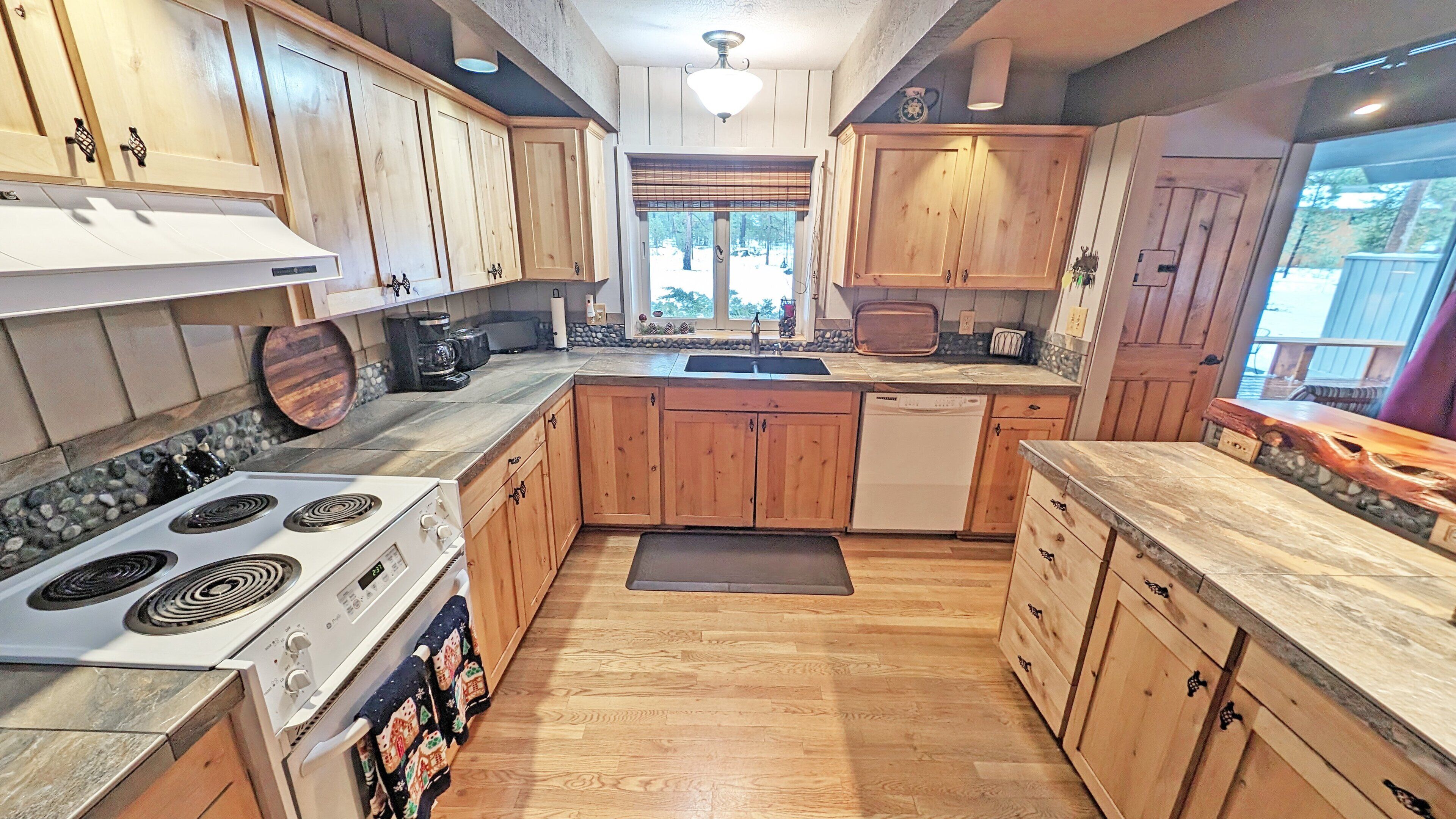 A kitchen with wooden cabinets, a stove, a dishwasher, and a window with a view of trees.