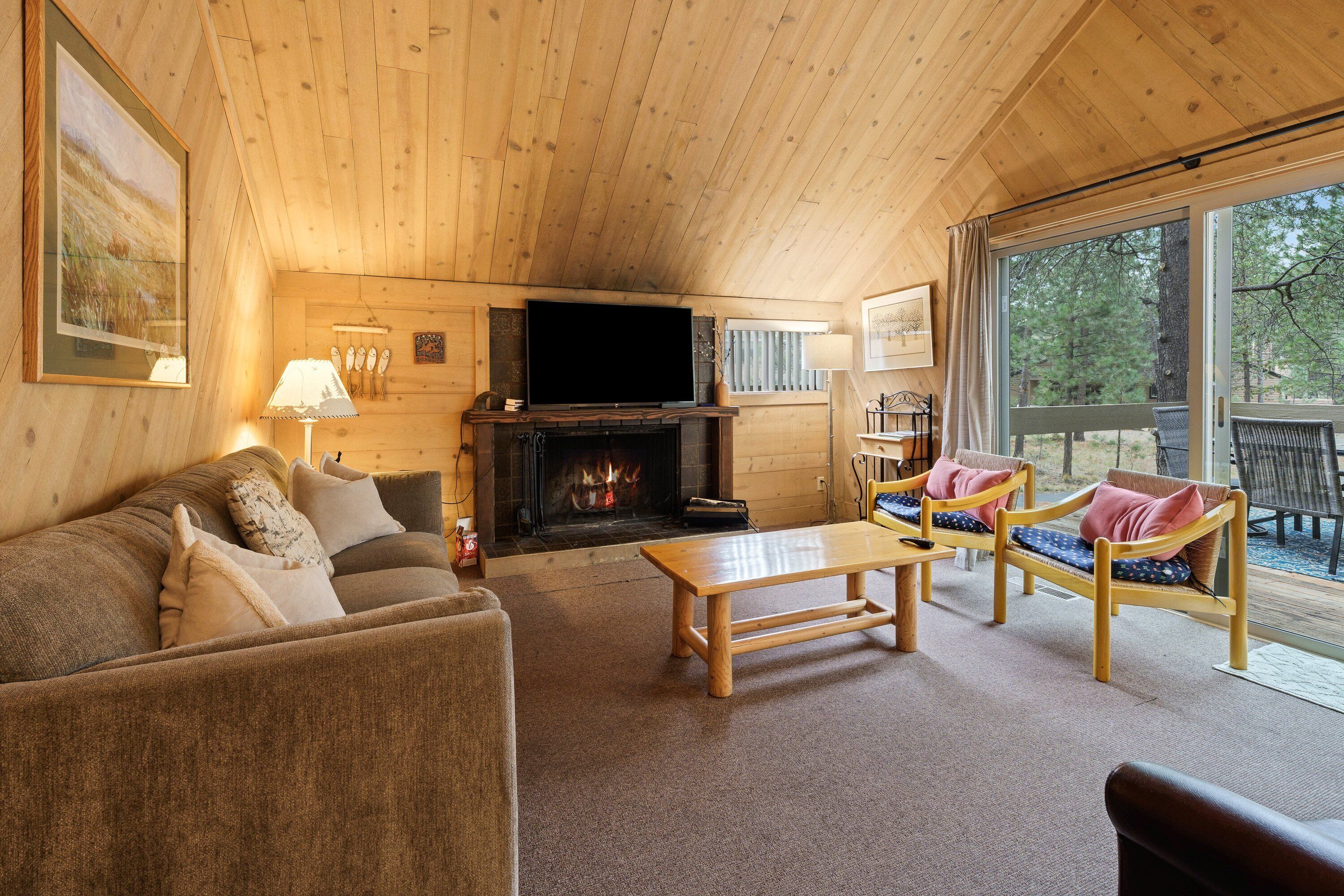 A cozy cabin interior with a wooden ceiling, a fireplace, a sofa, a coffee table, and a view through large windows.