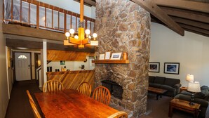 Un comedor con una mesa y sillas de madera, una chimenea de piedra, una cocina con armarios de madera y una sala de estar con sofá y lámpara.