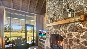 A stone fireplace with a fire, a wooden ceiling, a view of a grassy field, and a television. - Mt Bachelor Retreat - 221 (Bend)