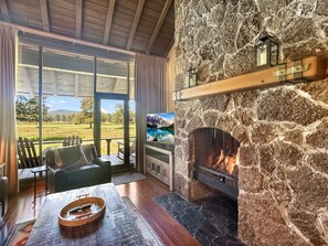 A stone fireplace with a fire, a wooden ceiling, a view of a grassy field, and a television. - Mt Bachelor Retreat - 221 (Bend)