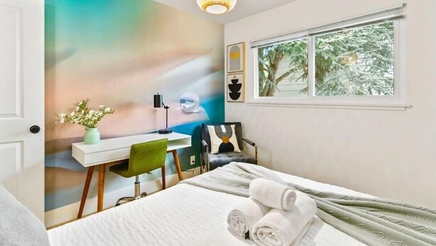 Ein Schlafzimmer mit einem Bett, einem Schreibtisch, einem Stuhl, einem Fenster und einer Wand mit einem farbenfrohen Wandgemälde.