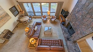 A spacious living room with a stone fireplace, a large window overlooking a deck, and a cozy seating area with a sofa and armchairs. - Goldfinch 3 (Bend)