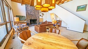 A spacious living room with a stone fireplace, wooden ceiling beams, and a large window. - Goldfinch 3 (Bend)