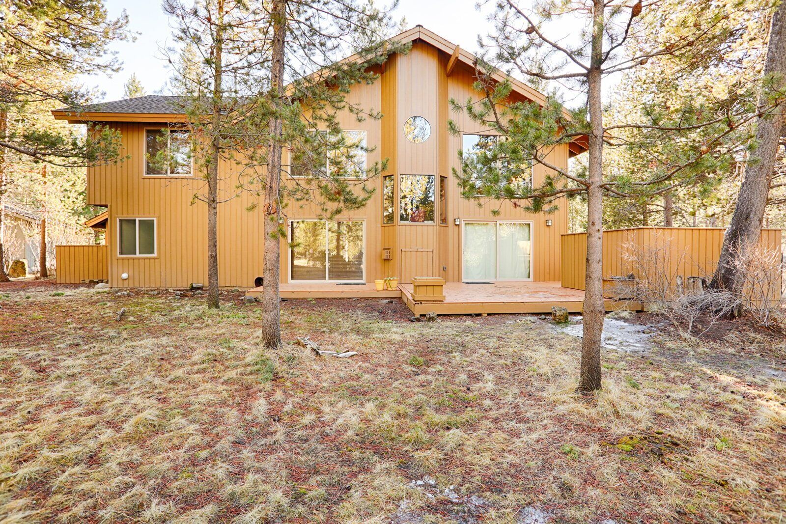 A wooden cabin with a deck, surrounded by trees and a grassy yard.