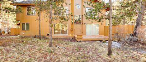 A wooden cabin with a deck, surrounded by trees and a grassy yard.