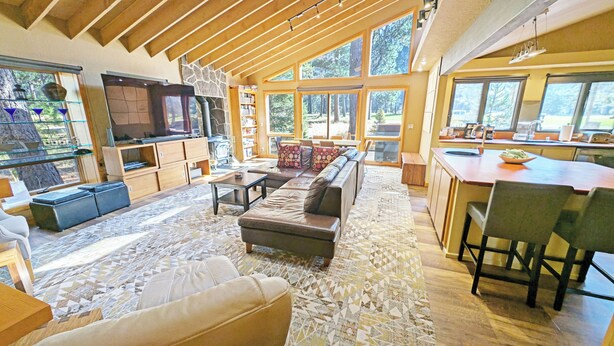 Amplia sala de estar con un sofá grande, una mesa de centro y una chimenea. Hay una zona de cocina con una mesa de comedor y sillas. La habitación tiene ventanas grandes que ofrecen luz natural y vistas al exterior.