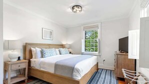 A bedroom with a wooden bed, a nightstand, a TV, and a window with a view of greenery. - Regatta Suite at The Royce Refined Newport Charm (Newport)