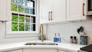 A modern kitchen with white cabinets, a stainless steel sink, and a window with a view of greenery. - Regatta Suite at The Royce Refined Newport Charm (Newport)