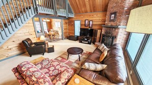 A cozy living room with a brown leather sofa, patterned ottoman, and a TV. A fireplace and wooden walls are visible. - Lava Top 1 (Bend)