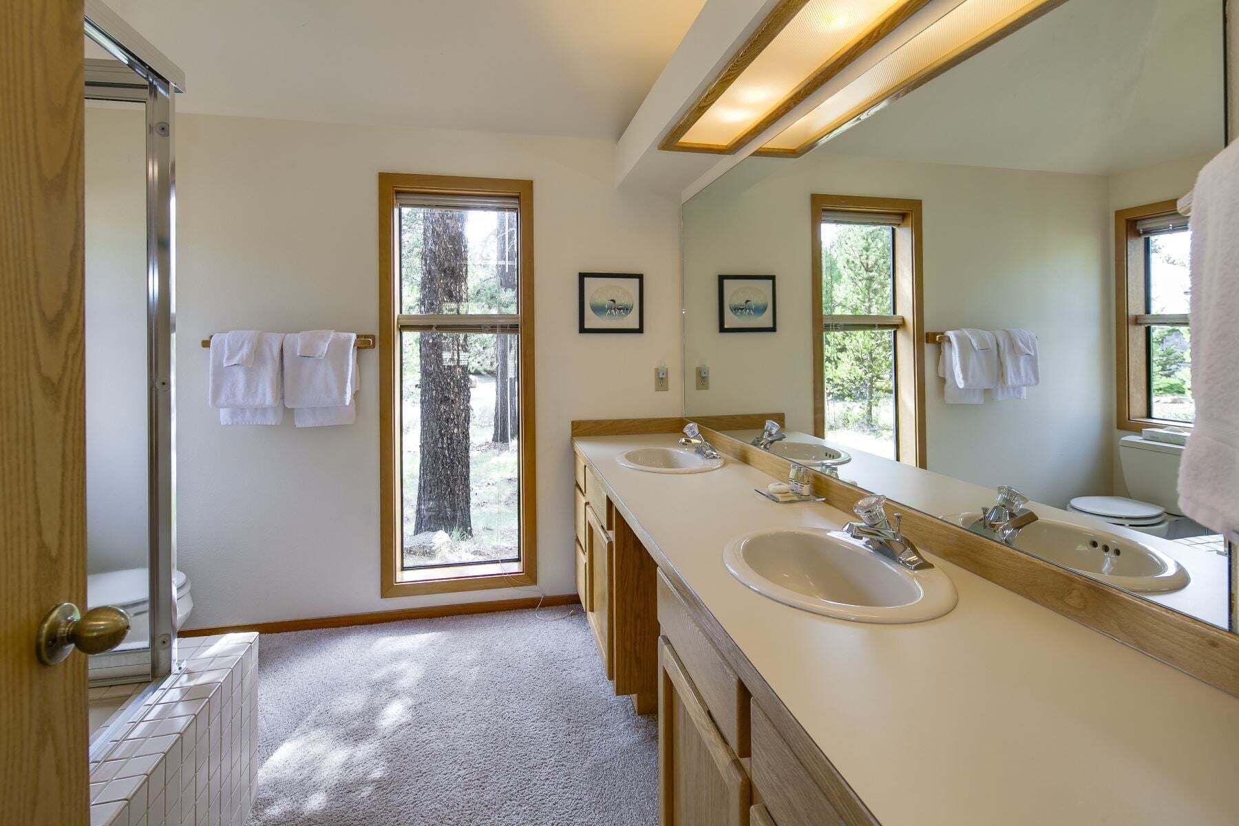 Une salle de bain avec deux lavabos, un grand miroir et une vue sur les arbres par les fenêtres.