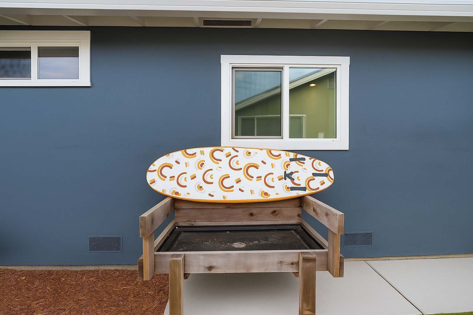 Un banco de madera con diseño de tabla de surf, ubicado frente a una casa de color azul y una ventana.