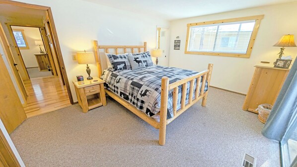 A bedroom with a wooden bed, a nightstand with a lamp, a window with blinds, and a door leading to another room.