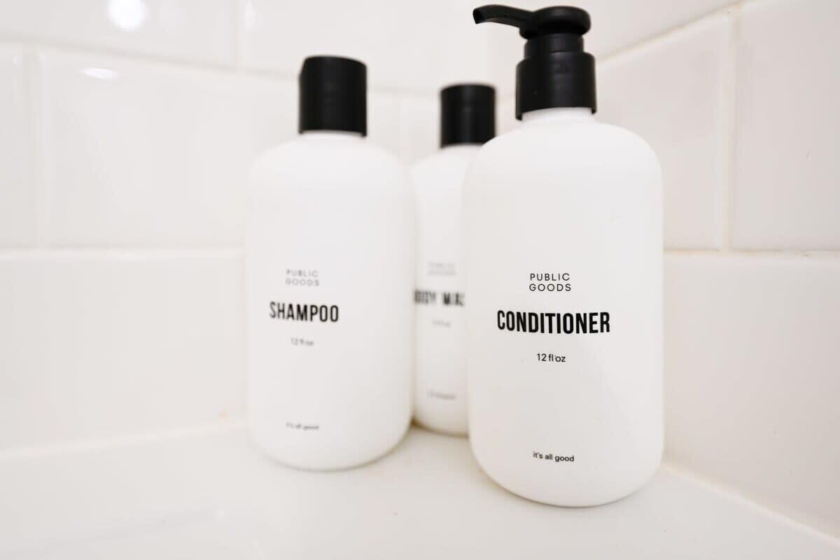 Tres botellas blancas de champú y acondicionador Public Goods sobre una superficie de azulejos blancos.