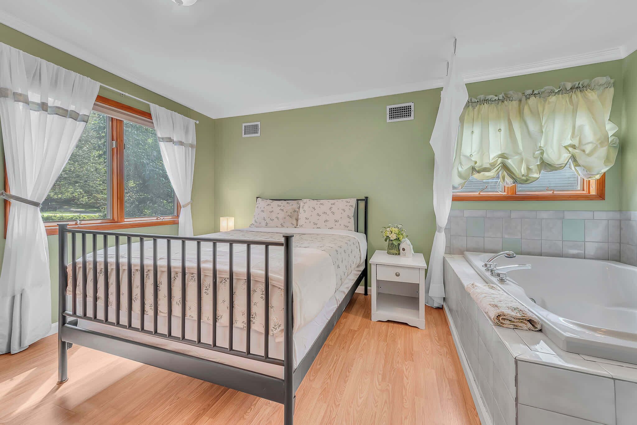 Un dormitorio con cama, mesita de noche, ventana con cortinas, baño con bañera y vistas a zonas verdes.
