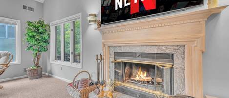 A cozy living room with a fireplace, a large TV mounted above it, and a comfortable seating area with a plant and a decorative dog statue.