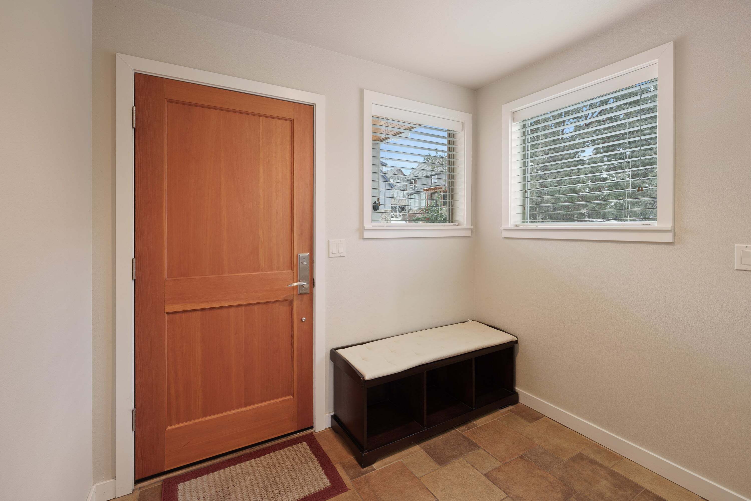 Un pasillo con una puerta de madera, un banco y dos ventanas con persianas.