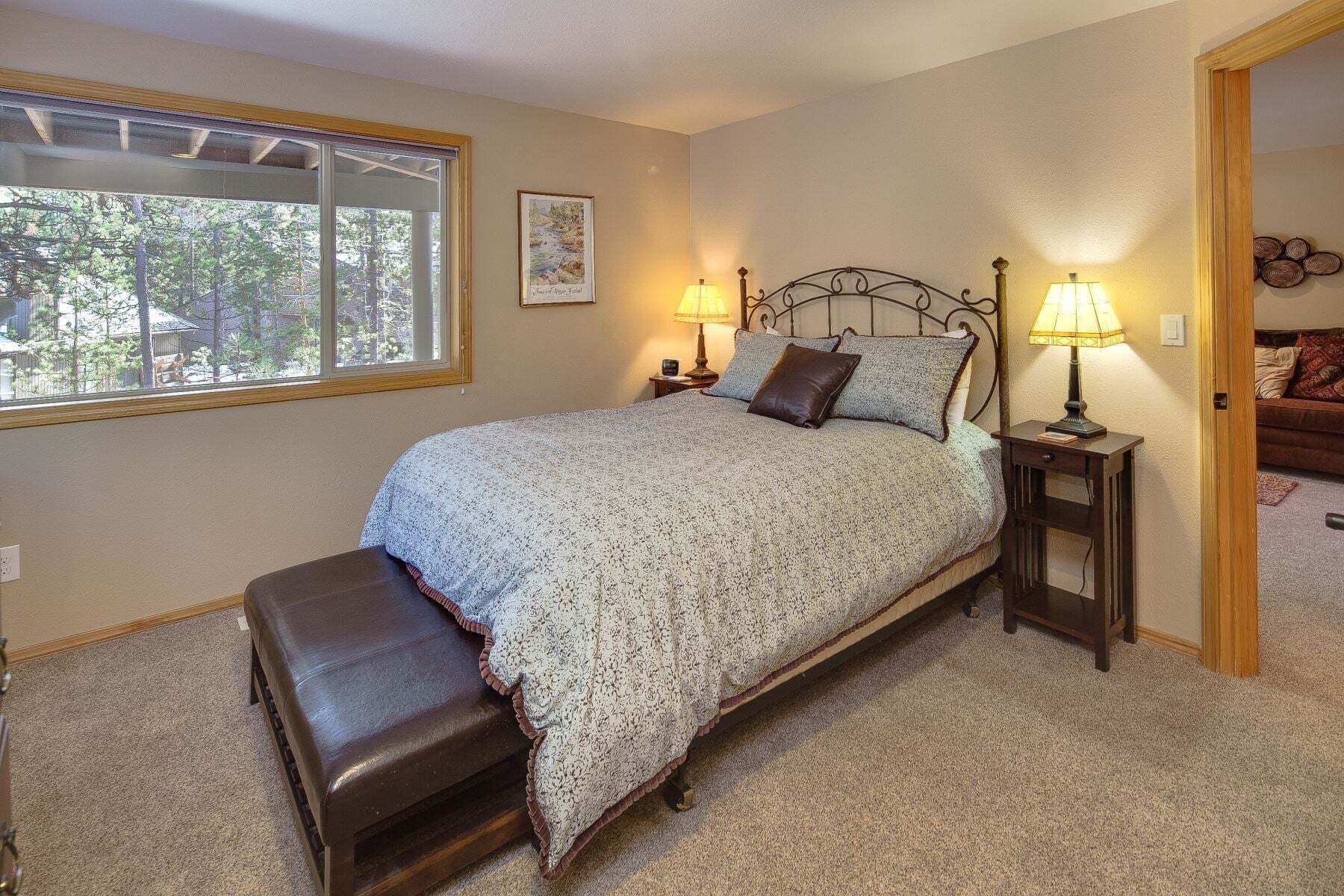 A bedroom with a bed, two bedside lamps, a window, a bench, and a door leading to another room.