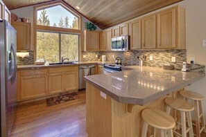 A kitchen with wooden cabinets, a stainless steel refrigerator, a microwave, a large island with stools, and a view of trees through the windows. - Lark 4 (Bend)