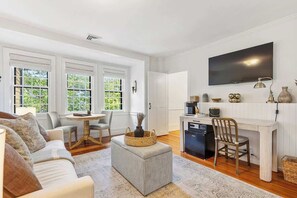 A spacious living room with a sofa, ottoman, and a dining area with a table and chairs. A kitchen area with a refrigerator, microwave, and a TV is visible in the background. - Driftwood Suite - Luxury Retreat in Newport (Newport)