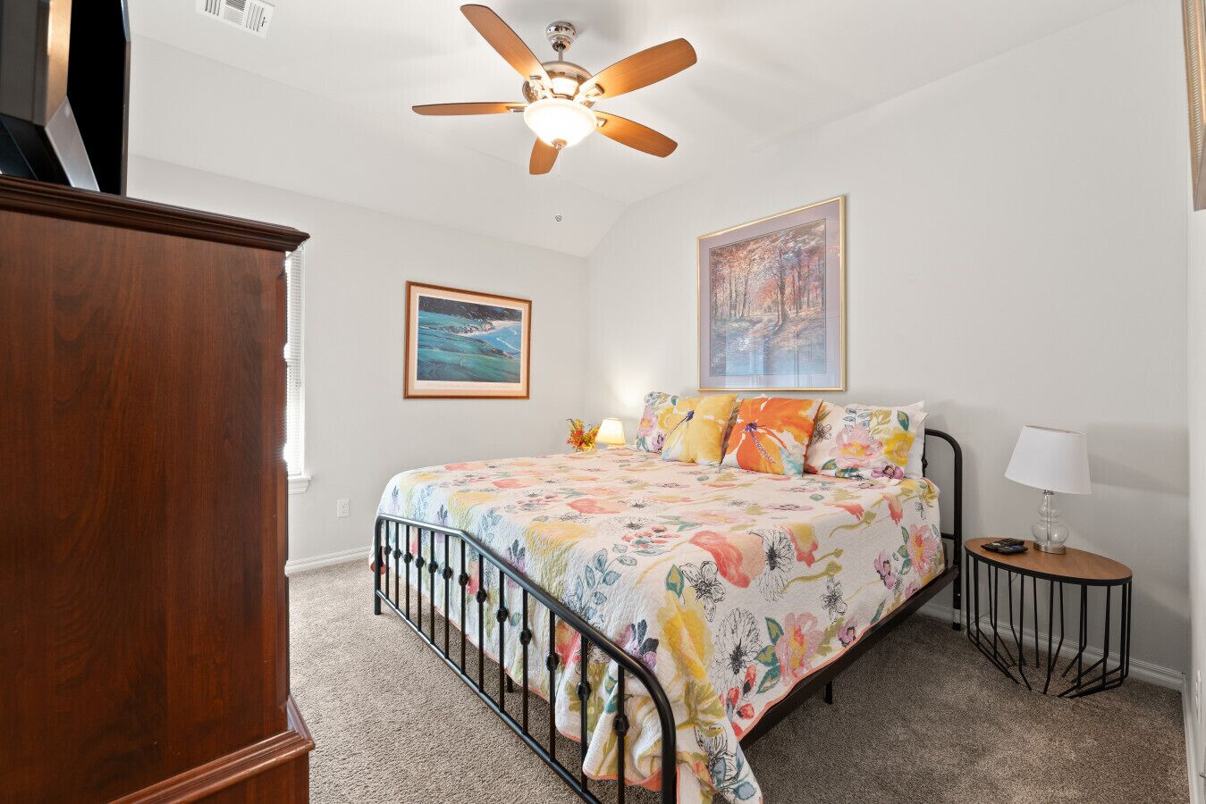 Une chambre à coucher comprenant un lit, un ventilateur de plafond, une table de chevet, une lampe et deux tableaux encadrés accrochés au mur.