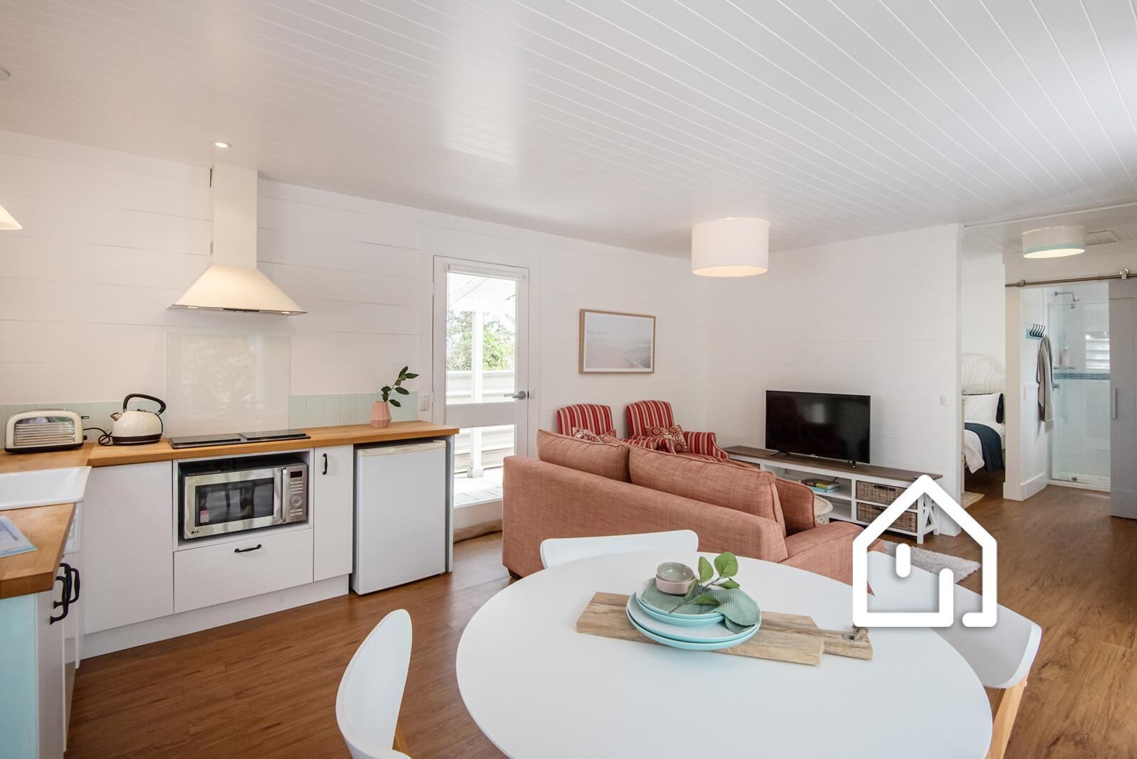A modern kitchen with a dining area, a living room with a sofa and TV, and a visible shower area.