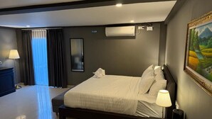 A bedroom with a large bed, a bedside table with a lamp, a wall-mounted air conditioner, and a painting on the wall. - Kooters Guesthouse Bali (Legian)