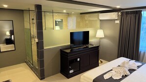 A hotel room with a glass-enclosed shower, a flat-screen TV on a dark dresser, a bed with neatly folded towels, and an air conditioning unit on the wall. - Kooters Guesthouse Bali (Legian)
