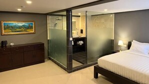 A modern hotel room with a bed, a dark wooden dresser, a glass partition, a bathroom with a mirror, and a painting on the wall. - Kooters Guesthouse Bali (Legian)