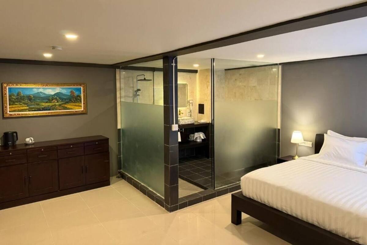 A modern hotel room with a bed, a dark wooden dresser, a glass partition, a bathroom with a mirror, and a painting on the wall.